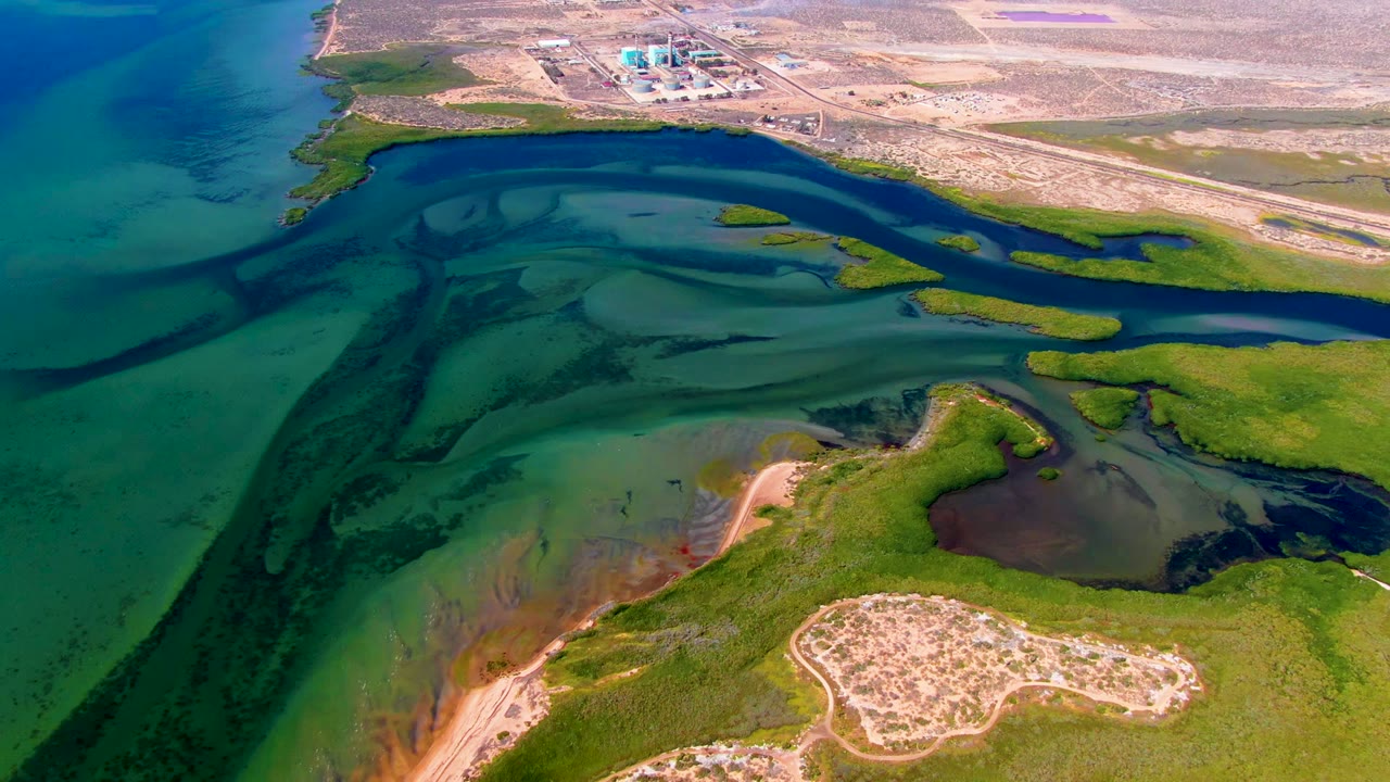 Opening aerial frame over San Carlos, Baja California Sur showing dark blue tidal channels, green marsh, tan sandbars, dry shoreline, distant industrial structures, and no visible people.