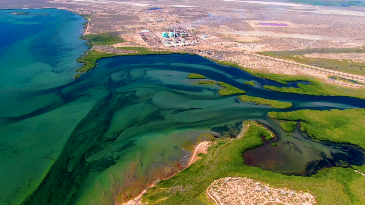 Aerial frame in San Carlos, Baja California Sur, with darker tidal water crossing shallow green-blue flats, marsh islands, dry inland terrain, distant structures, and no visible people.