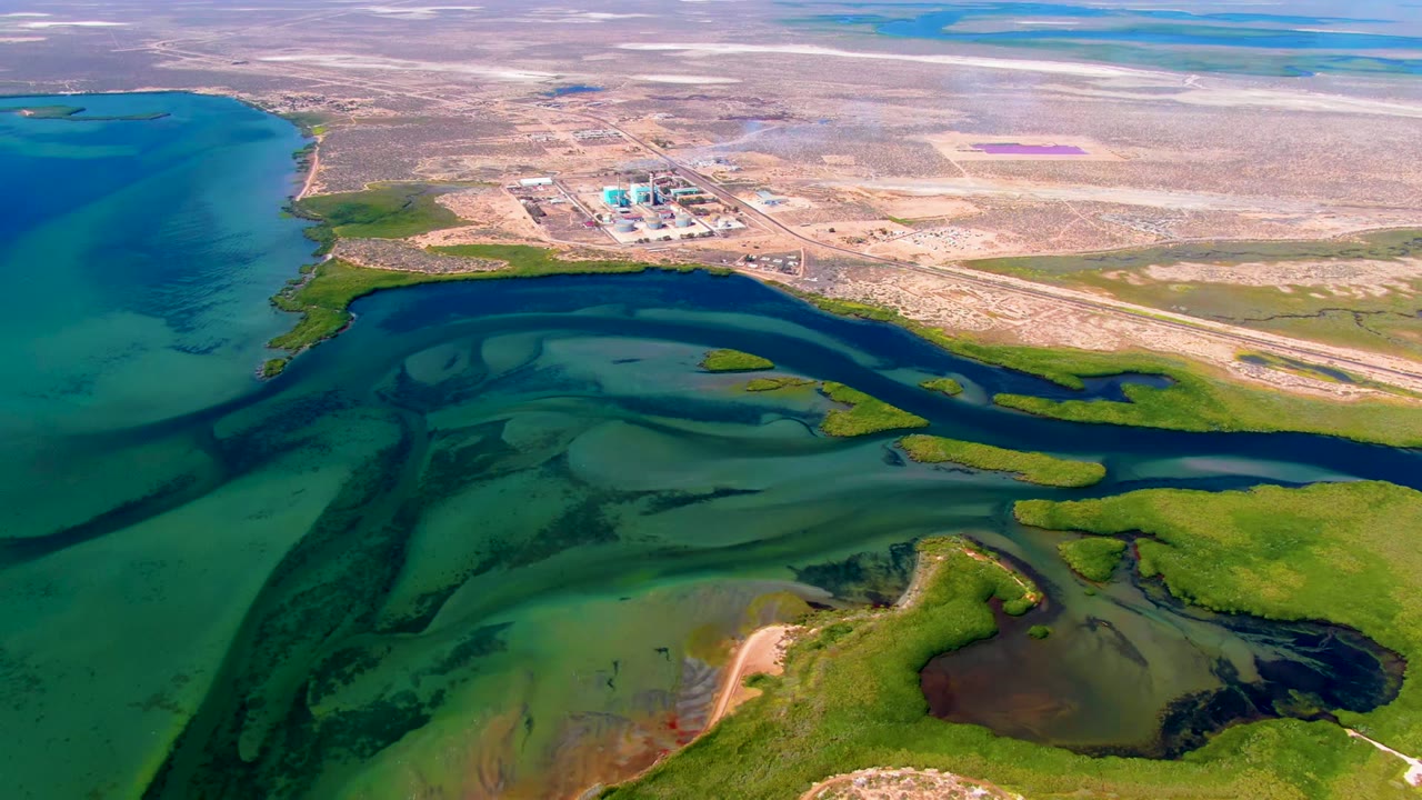 Aerial frame over San Carlos, Baja California Sur showing turquoise coastal water, dark wetland channels, green marsh vegetation, pale desert land, distant industrial structures, and no visible people.