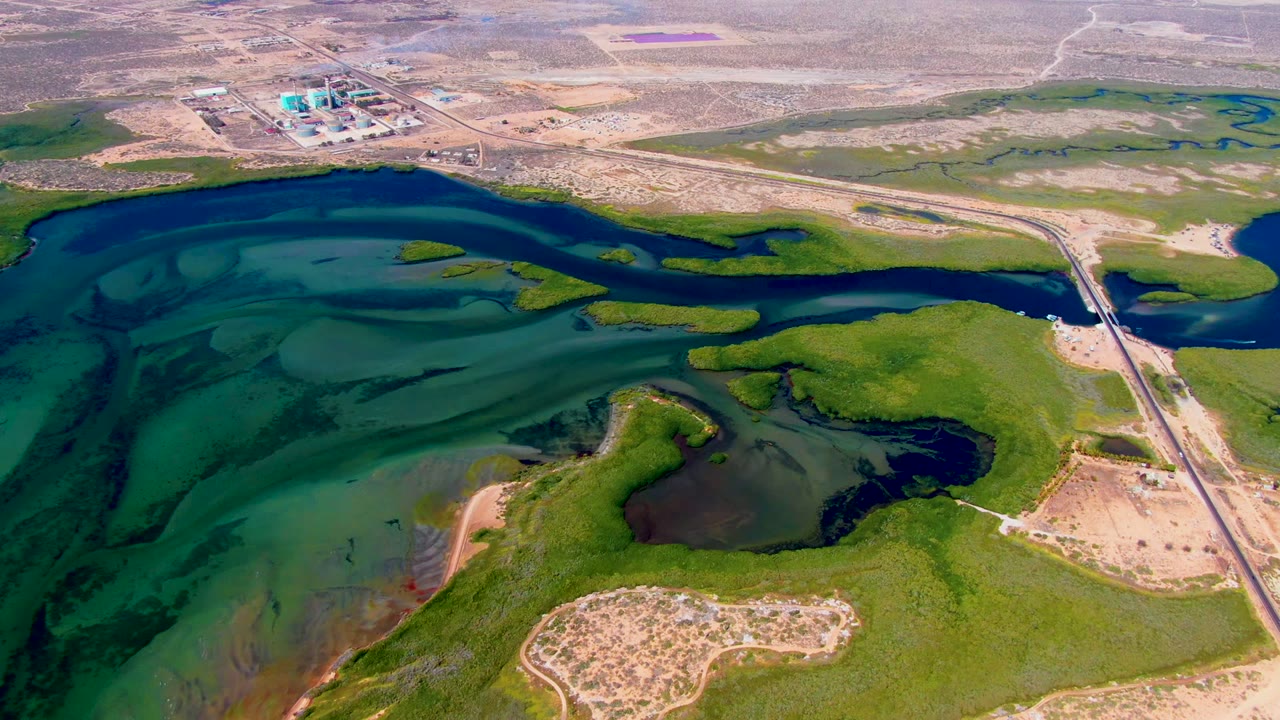 Aerial view over San Carlos, Baja California Sur showing the bridge crossing beside a dark blue channel, green wetland patches, tan desert flats, a small vehicle, and no visible people.