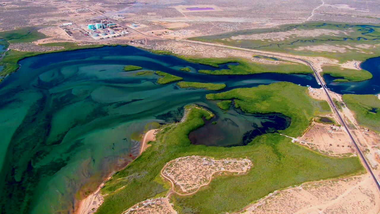 Wide drone view of reservoir water, green shoreline, road, and industrial buildings in the distance.