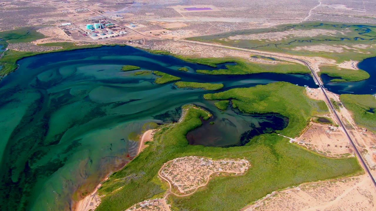 Third frame from the San Carlos aerial shows layered blue and green water, low wetland vegetation, sandy banks, the roadside area to the right, distant industrial structures, and no visible people.