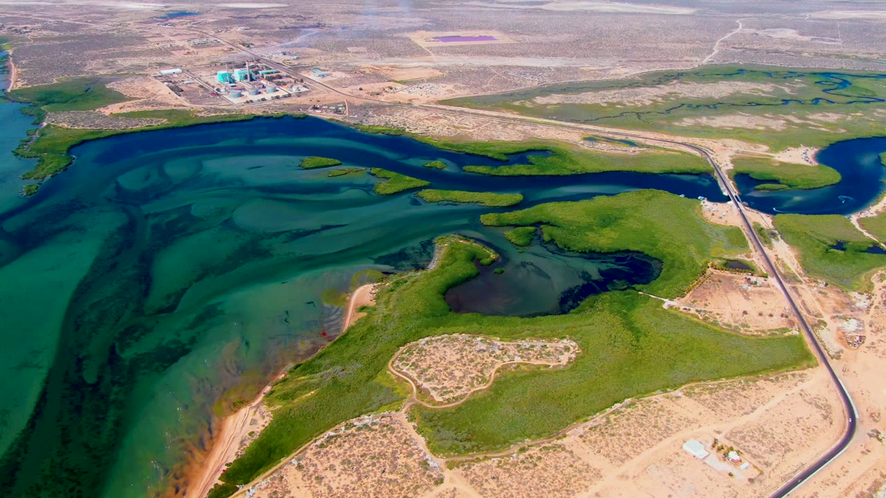 Opening aerial frame over San Carlos, Baja California Sur reservoir with blue-green water at left, green shoreline in the middle, tan dry terrain, a road and bridge at right, distant industrial buildings, and no visible people.