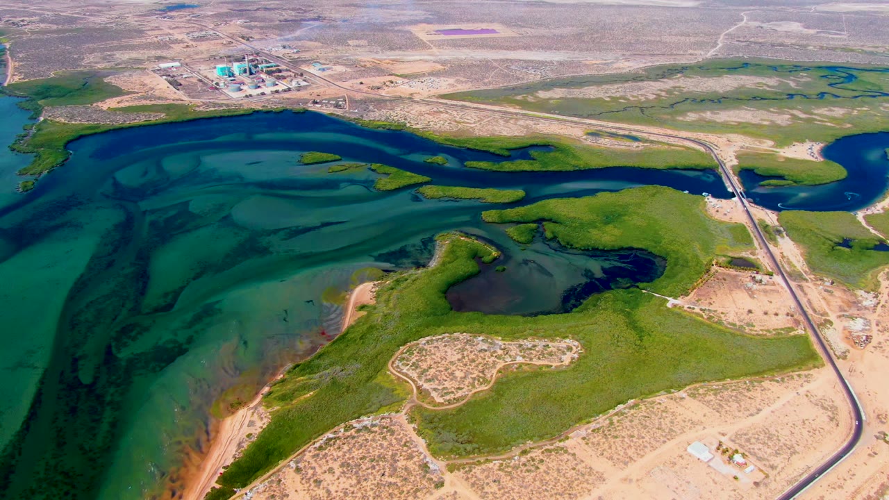 Aerial reservoir scene with shallow water channels, green land, road bridge, and industrial buildings beyond dry terrain.