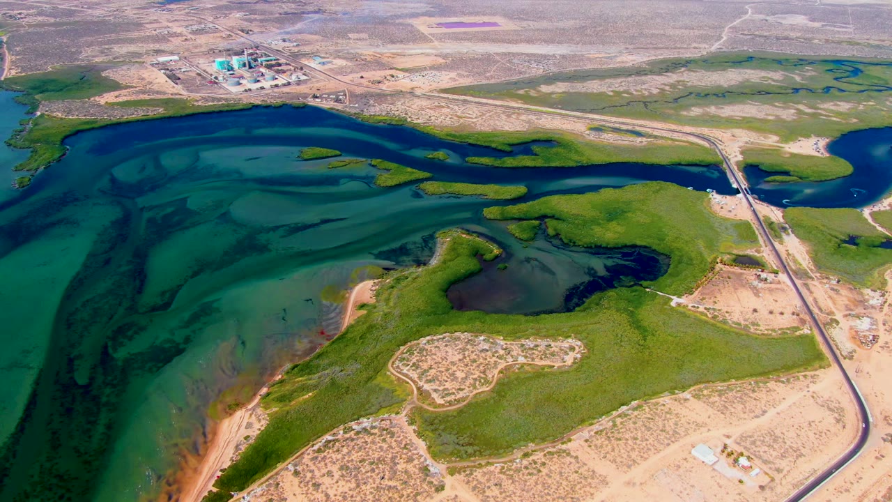 Third frame over San Carlos, Baja California Sur with broad turquoise reservoir shallows at left, narrow green islands and banks across the center, a paved road and bridge at right, distant industrial structures, and no visible people.
