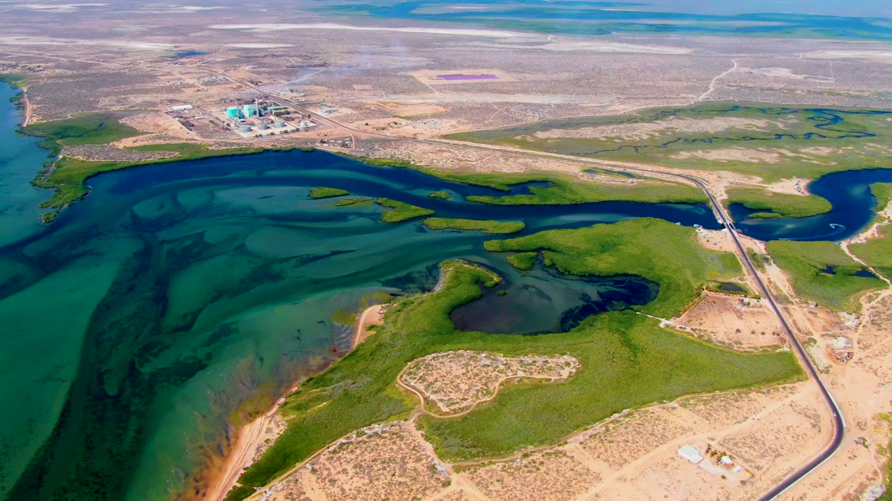 Aerial reservoir view with dark blue channels, green algae mats, sandy banks, and road at right.