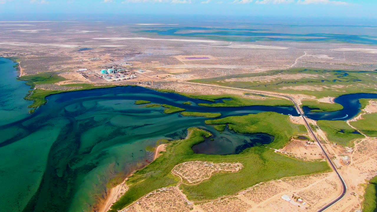 Drone view of shallow water, marsh channels, roadway, and dark spill areas near the shore.