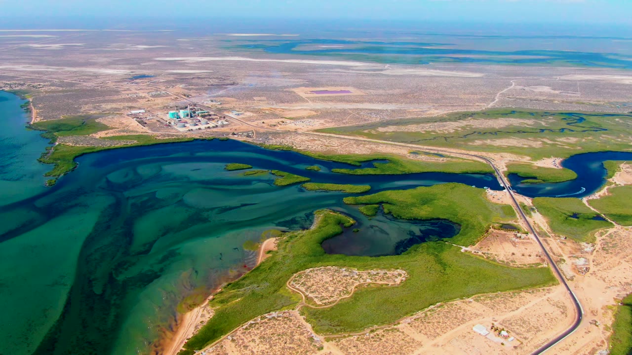 At 0:02, dark spill streaks remain visible across the teal shallows off San Carlos, Baja California Sur, with marsh green, a pale road, distant structures, and no visible people.