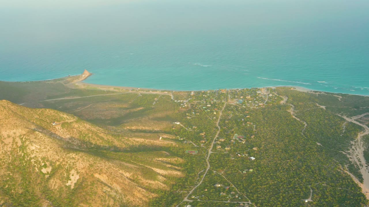 Opening aerial frame over Cabo Pulmo, Baja California Sur, with brown hillside foreground, green settlement areas, sandy shore, turquoise sea, and no visible people.