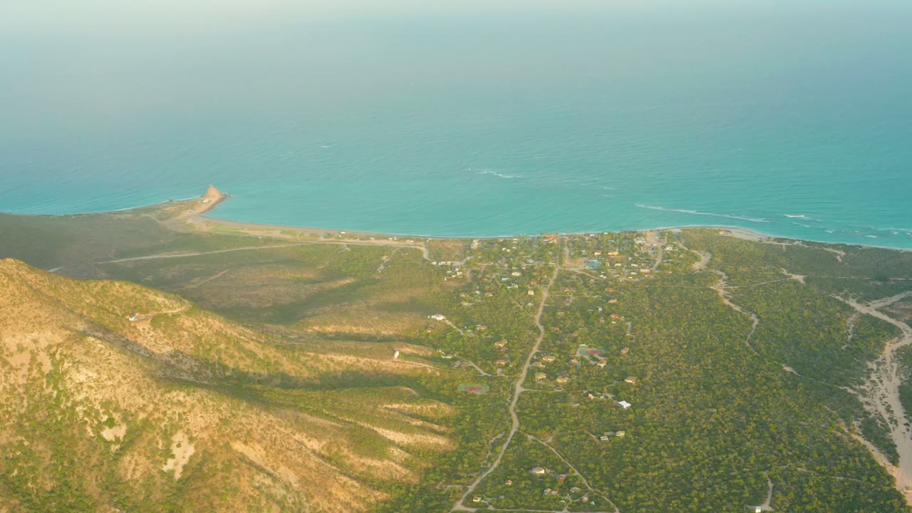 Wide aerial coastline with hillside roads, small buildings, beach, and blue-green sea.