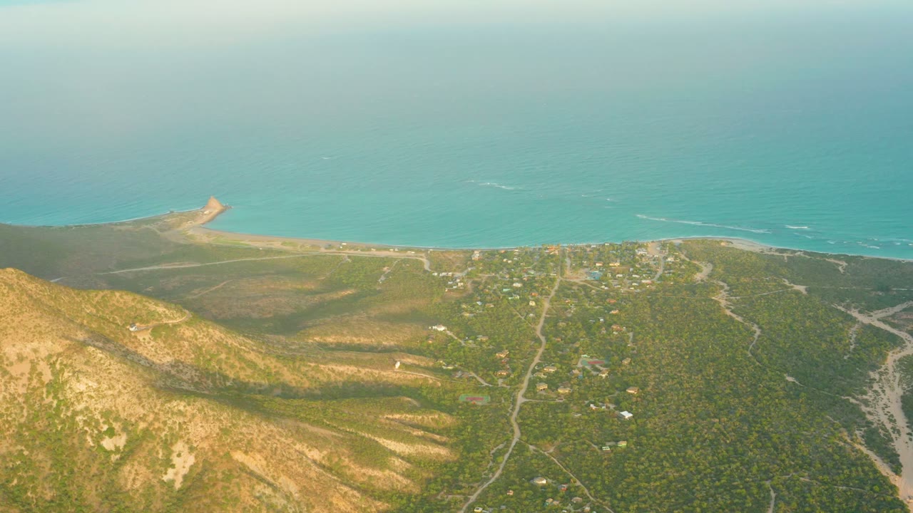 Cabo Pulmo aerial frame with sunlit brown hills in the lower frame, a patchwork of green vegetation and scattered structures in the center, turquoise ocean above, and no visible people.