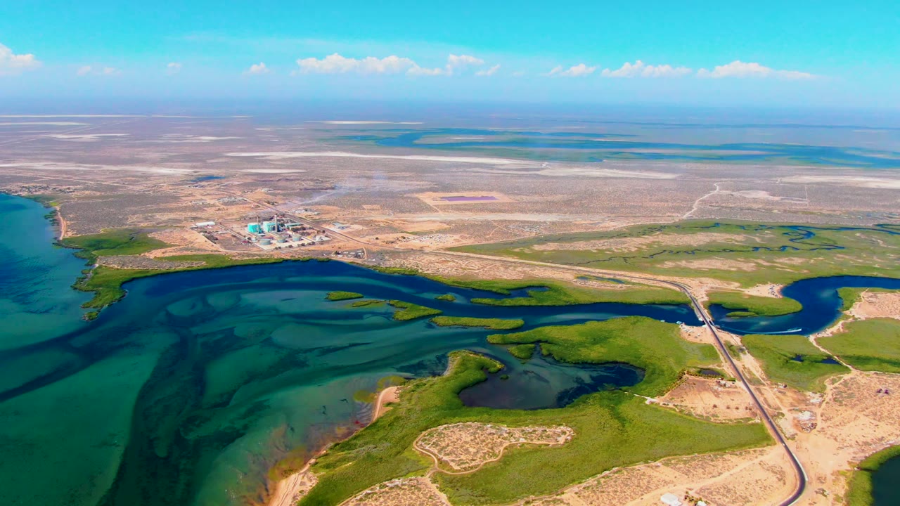 Aerial frame from San Carlos, Baja California Sur showing a factory smoke plume beyond turquoise water, green wetlands, tan desert flats, and a road crossing the shoreline, with no visible people.