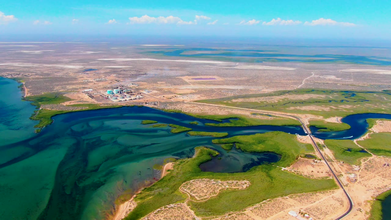 Aerial frame in San Carlos, Baja California Sur showing a narrow road and bridge near the water, green marshy areas in the foreground, and a smoke plume rising from the factory in the distance with no visible people.