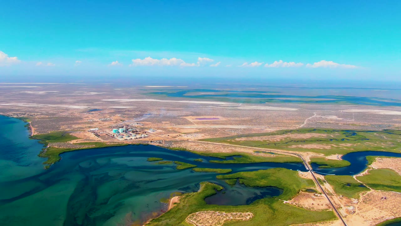 Aerial view over San Carlos, Baja California Sur, with turquoise coastal water in the foreground, green wetland channels, dry flats, distant buildings, and no visible people.