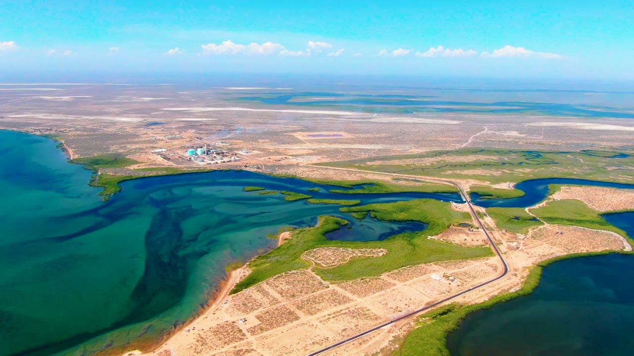 Aerial frame over San Carlos, Baja California Sur showing turquoise coastal water at left, green wetland shapes, a road or runway crossing the shoreline, distant cleared land, and no visible people.