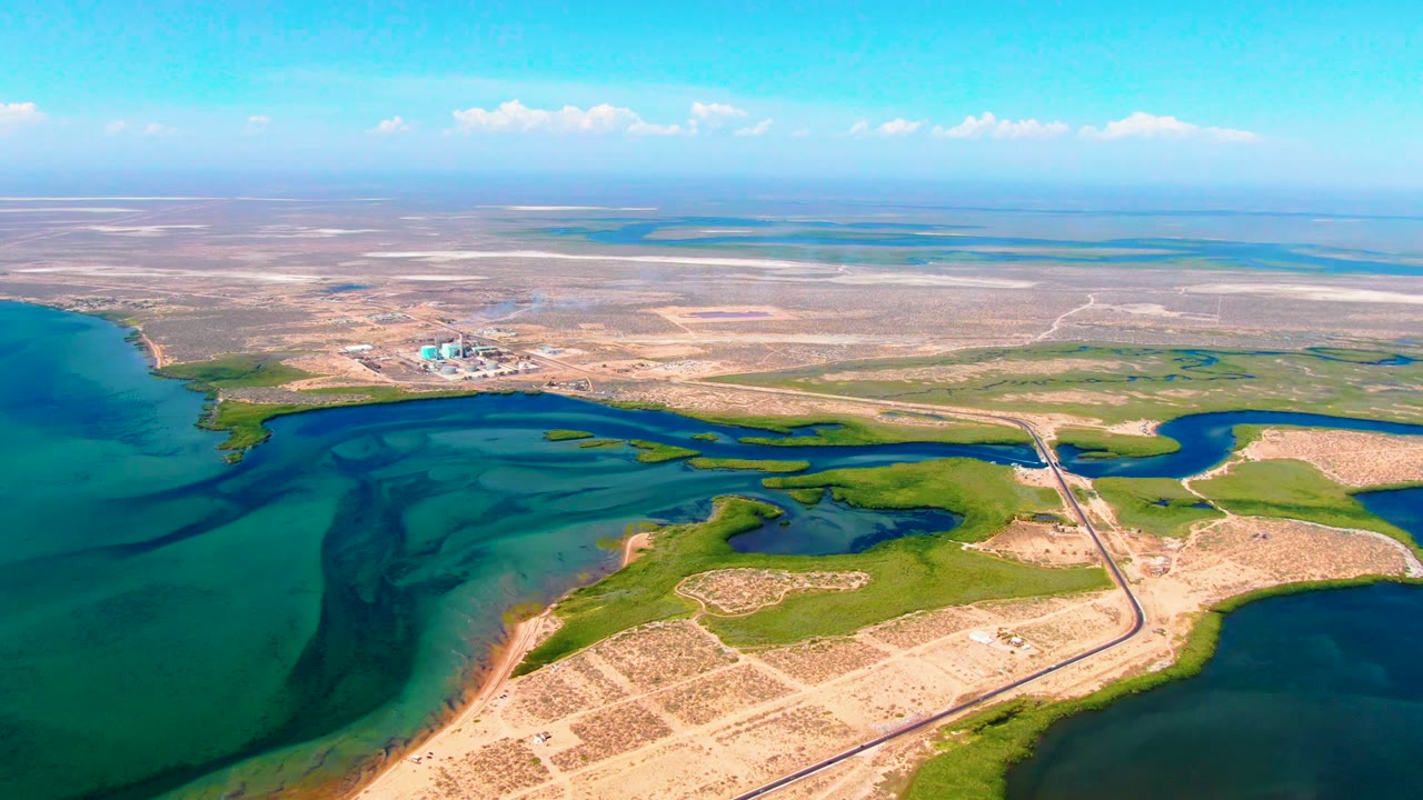 Aerial coastline with blue-green water, marsh islands, sandy tracks, and construction in the distance.