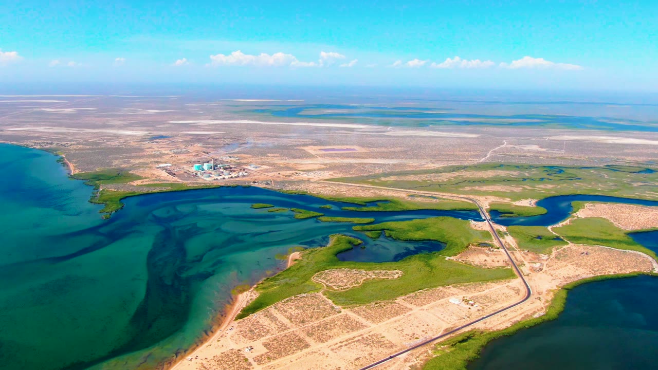 Aerial frame from San Carlos, Baja California Sur showing shallow-looking coastal channels, bright green land patches, tan cleared plots, a roadway crossing the frame, a background construction area, and no visible people.
