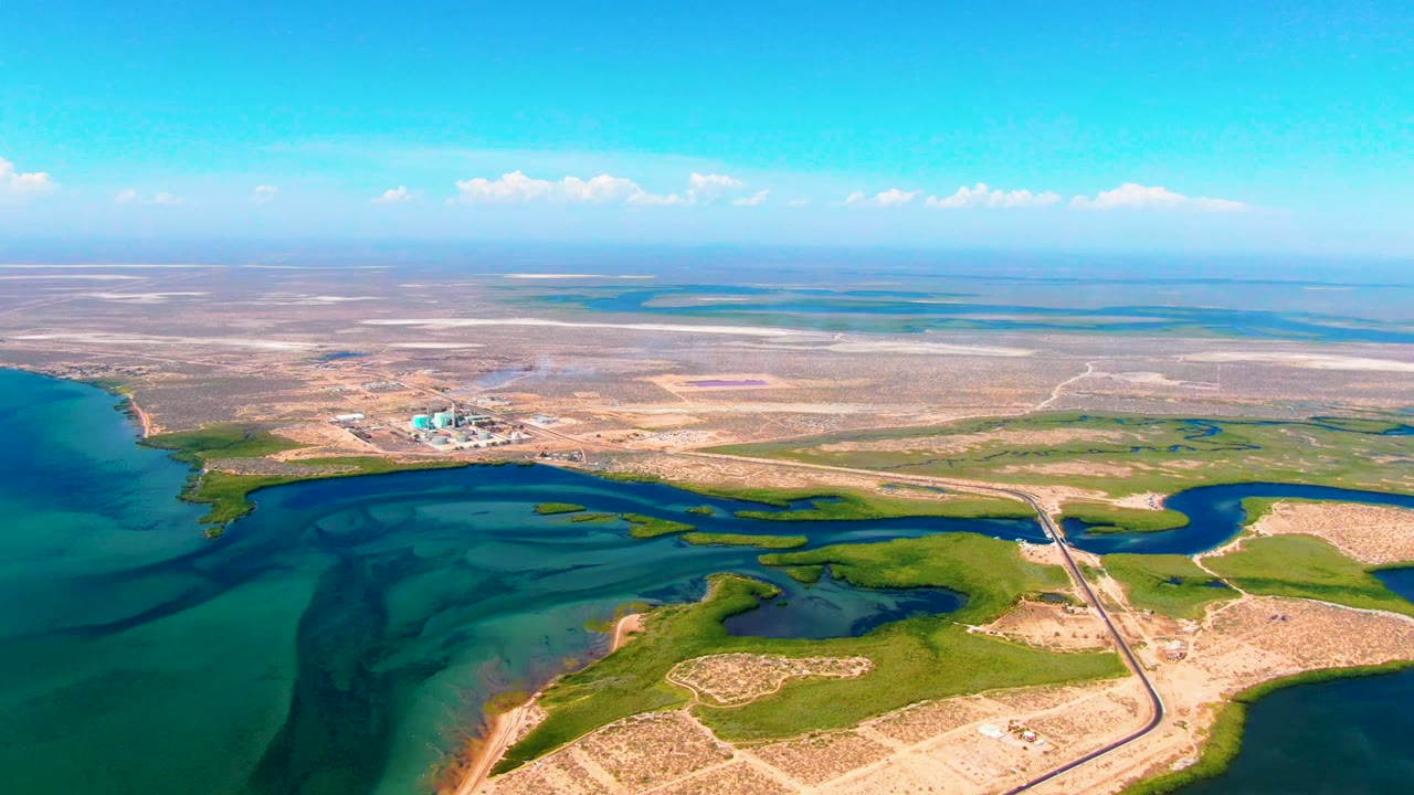 Wide drone view over San Carlos, Baja California Sur, with turquoise water in the foreground, green wetland channels, a road crossing the shore, a distant industrial plant, faint smoke, and no visible people.