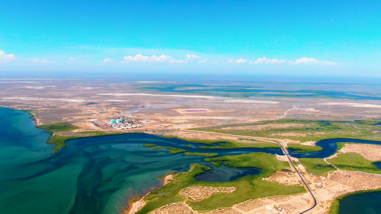 San Carlos aerial view with darker blue water at left, green wetland patches across the lower frame, a straight road and crossing near the right, distant industrial buildings, smoke, and no visible people.