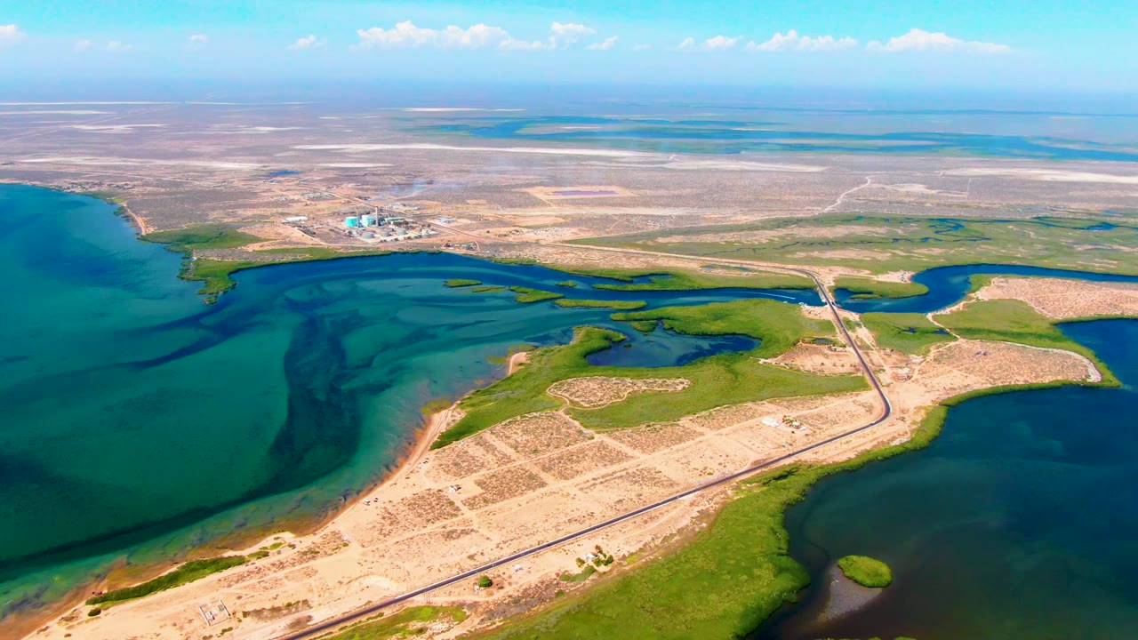Aerial frame from San Carlos, Baja California Sur showing a narrow causeway crossing between blue-green water, sandy flats, green patches, and a distant horizon with no visible people.