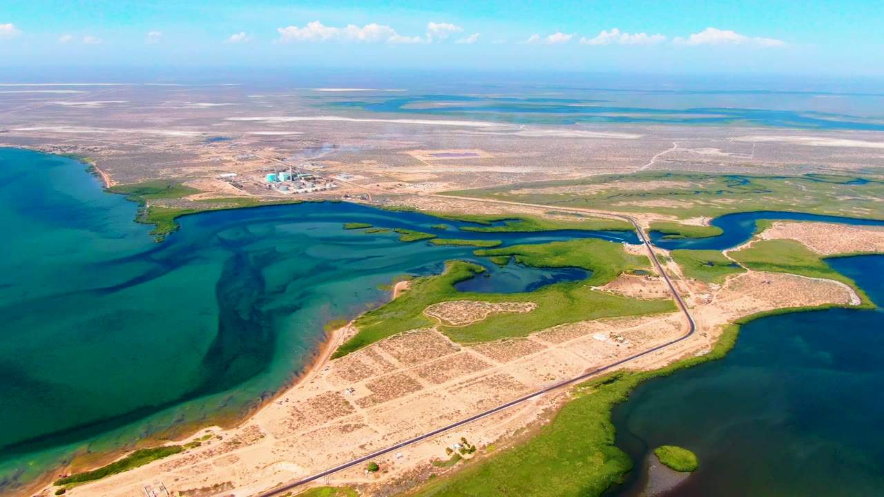 Wide drone frame over San Carlos showing turquoise water on the left, darker water at right, the causeway through the center, dry open terrain beyond, and no visible people.