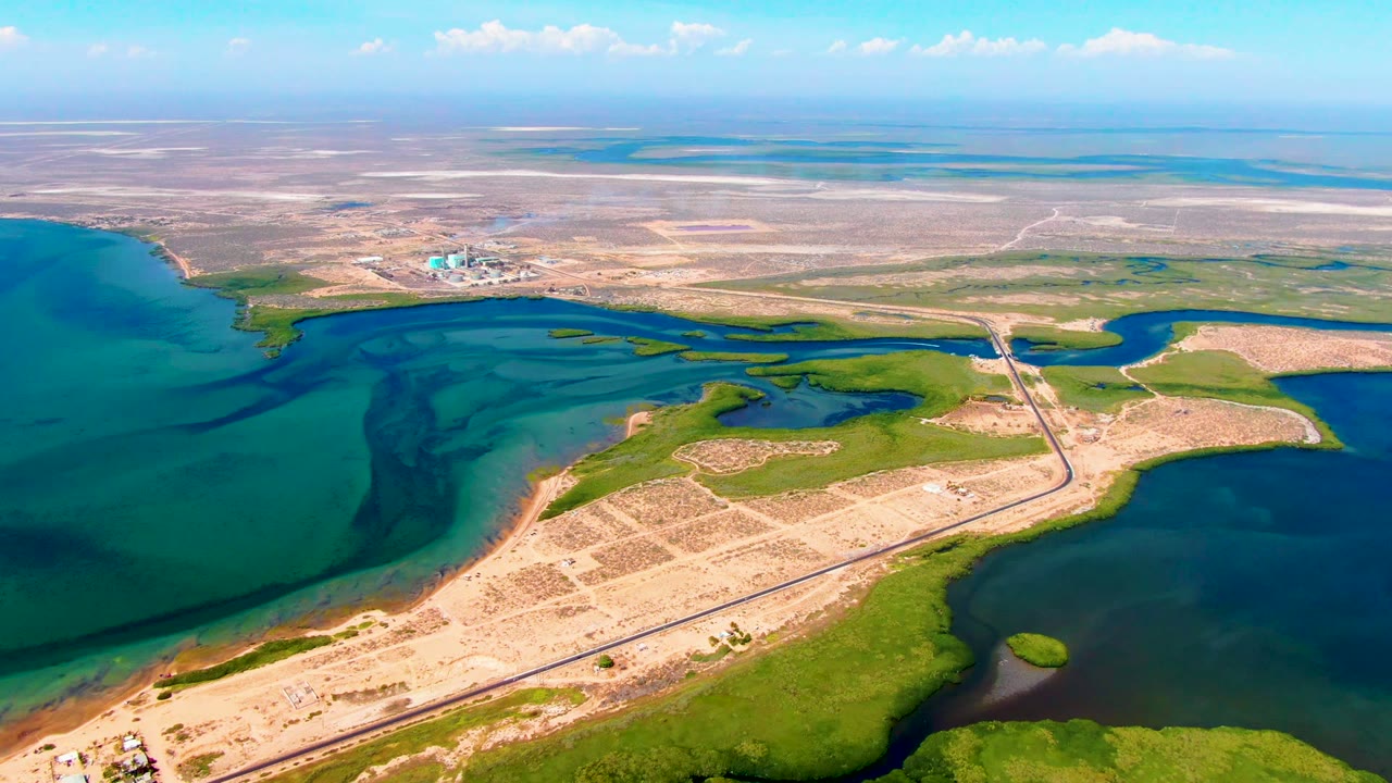 Opening aerial frame over San Carlos, Baja California Sur, showing a coastal road along sandy land, blue-green water, marshy patches, distant desert flats, and no visible people.