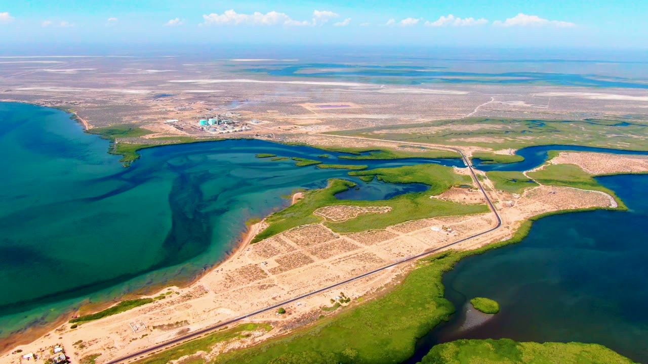 Drone view of a road crossing sandy land between water and green wetland areas.