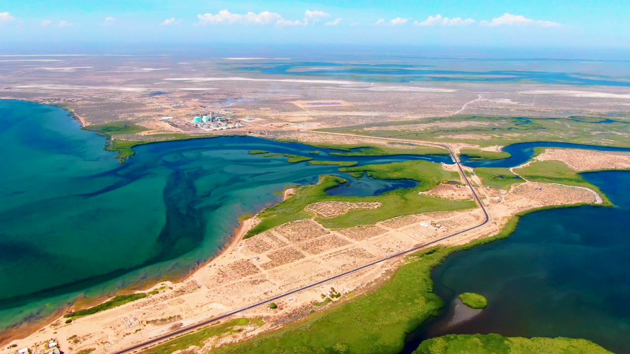 At 0:02, the view over San Carlos shows turquoise and dark blue water, irregular green channels, a pale road across sandy ground, a small distant settlement area, and no visible people.