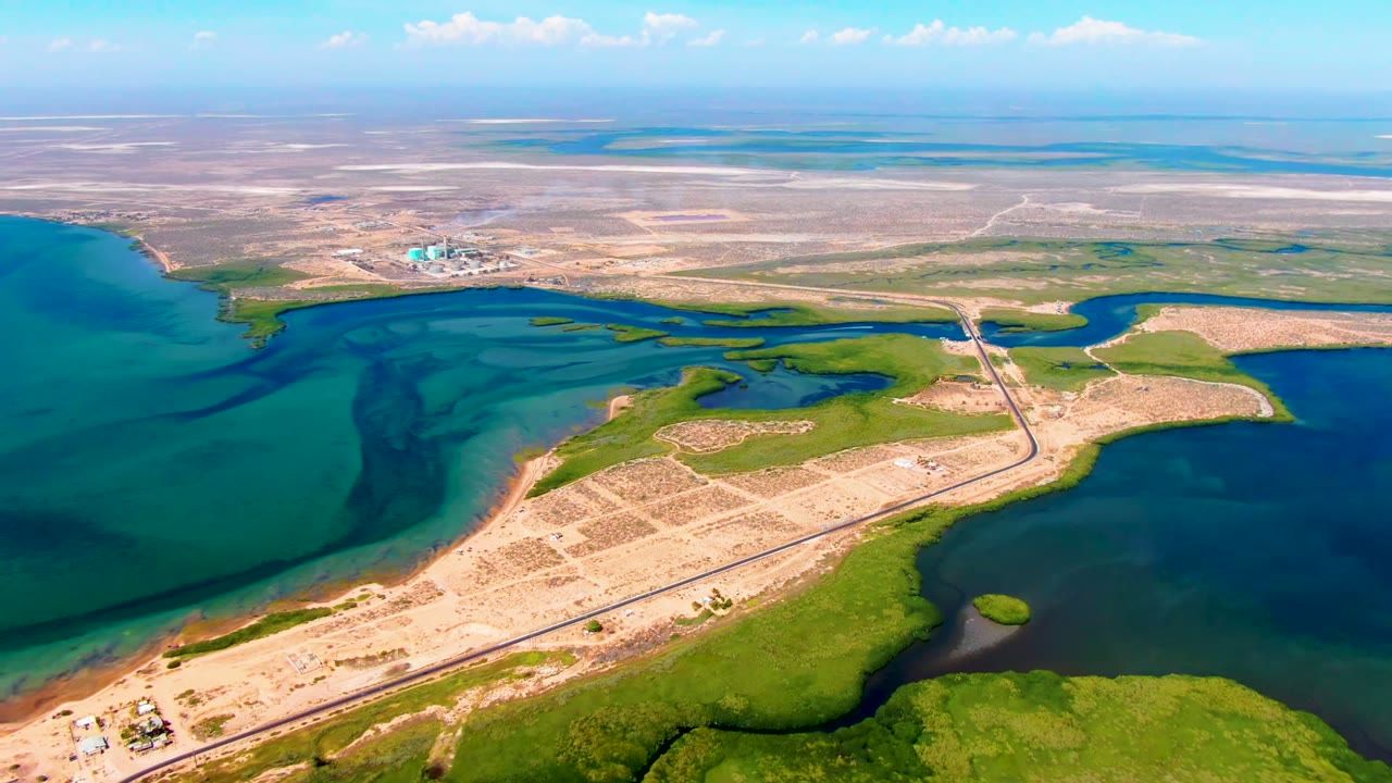 Wide aerial view in San Carlos, Baja California Sur showing blue coastal water, green wetlands, a straight road with bridge, sandy open land, distant buildings, and no visible people at the start of the clip.