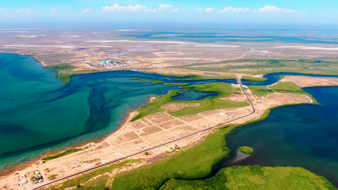 Wide San Carlos, Baja California Sur aerial view showing the road crossing a bridge near green wetland edges, blue water on both sides, open tan terrain, a pale horizon, and no visible people.