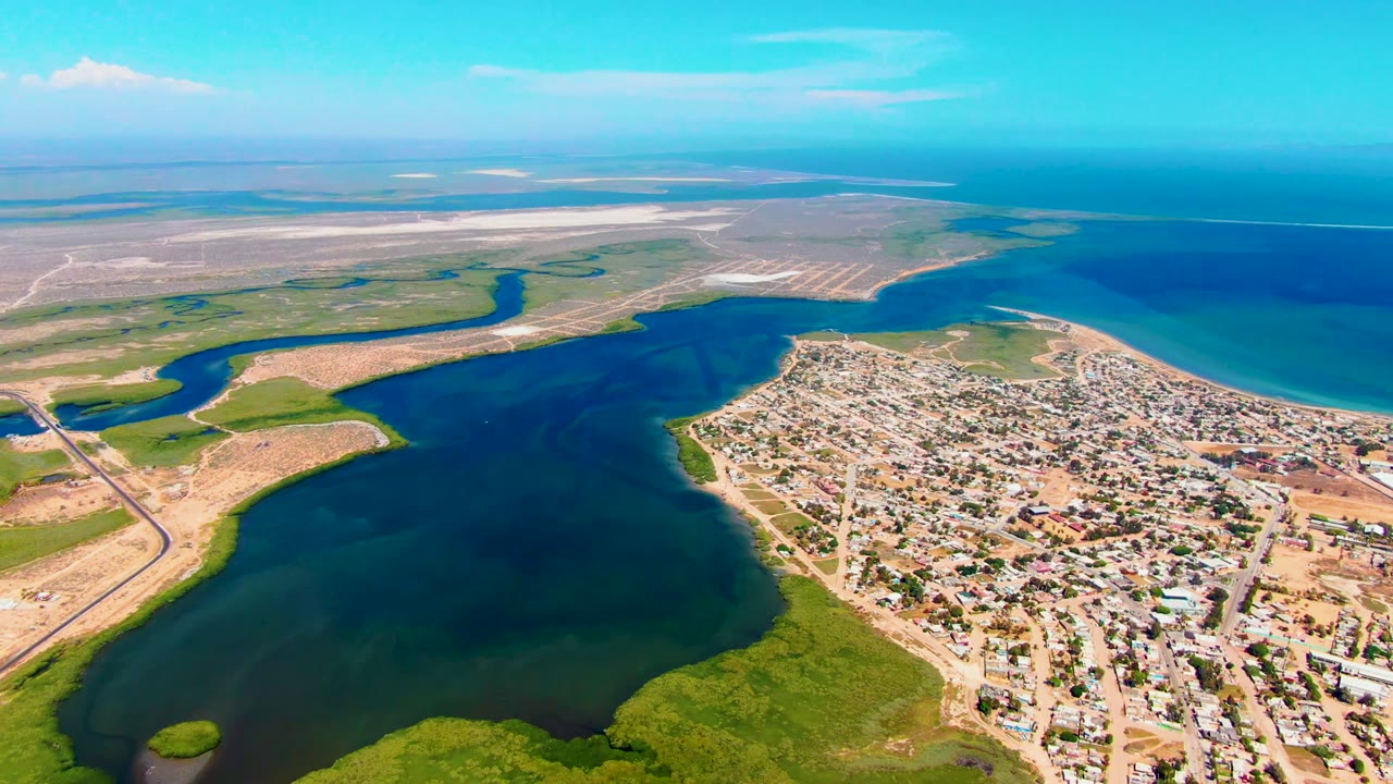 Opening aerial frame over San Carlos, Baja California Sur, showing a dark blue inlet, green wetland shoreline, sandy flats, dense town blocks, turquoise ocean, and no visible people.