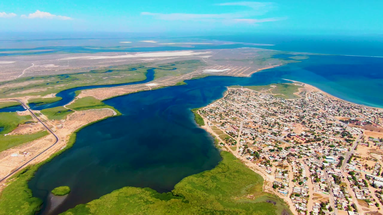 Mid-clip aerial view in San Carlos, Baja California Sur, showing the inlet pressed against a built-up town, green wetland foreground, pale exposed shoreline, open ocean, and no visible people.