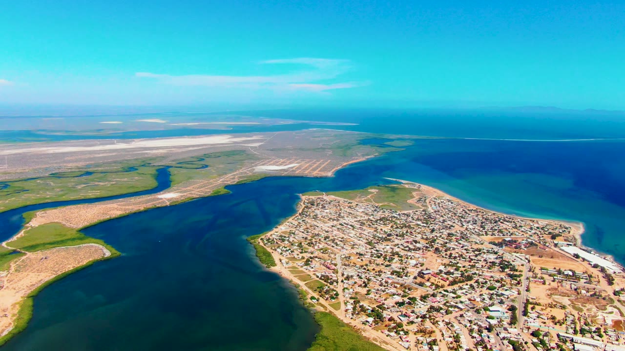 Aerial view of San Carlos, Baja California Sur, with a compact coastal town below, dark inlet water at left and center, sandy flats beyond, and no visible people.