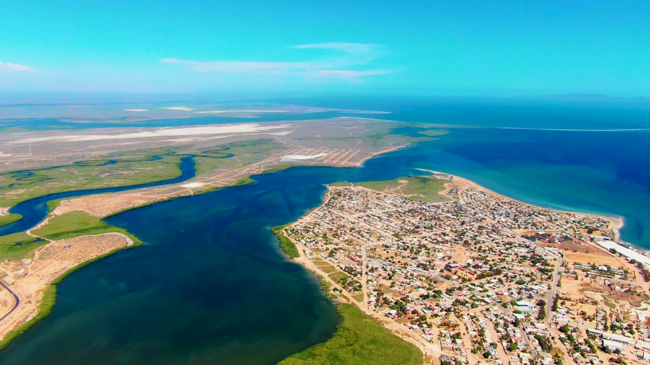 Wide aerial frame of San Carlos with the town on a sandy peninsula, blue-green water wrapping around it, marsh vegetation in the foreground, and no visible people.