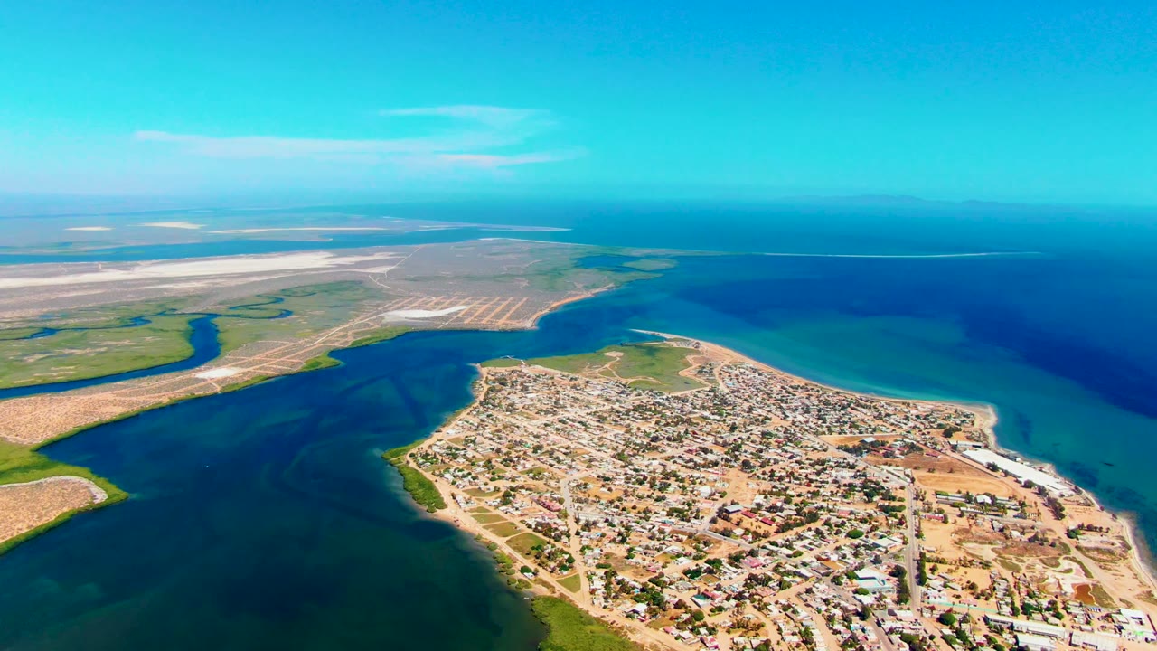 Aerial view over San Carlos, Baja California Sur, with residential blocks in the lower frame, a dark water channel at left, turquoise ocean at right, sandy flats beyond, and no visible people.