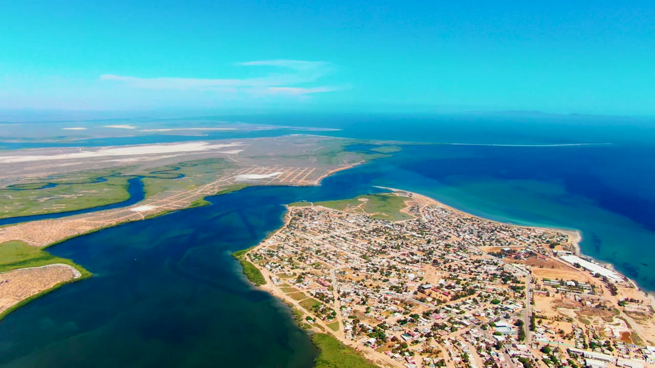 San Carlos, Baja California Sur appears from above with dense low buildings near the shore, a dark waterway wrapping the left side, bright ocean water to the right, and no visible people.