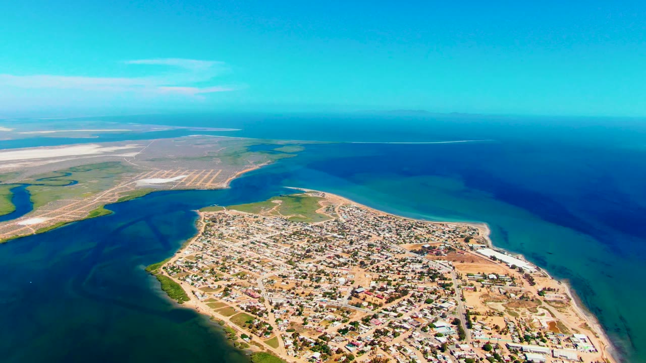 Opening aerial frame over San Carlos, Baja California Sur, with the town on a peninsula, turquoise shallows, deep blue ocean, pale coastal flats, and no visible people.