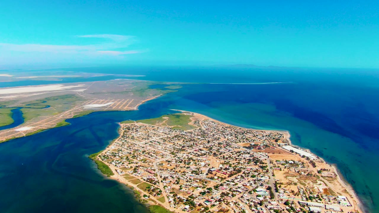 High aerial over a peninsula town with shallow turquoise water and open sea beyond.