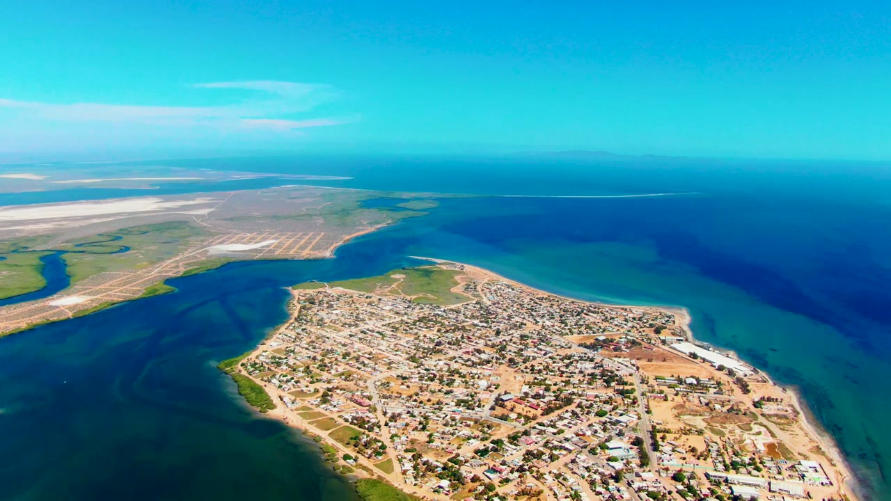 Aerial view of San Carlos with the settlement across the lower peninsula, light sandy lots, green coastal edges, broad blue water, and no visible people.