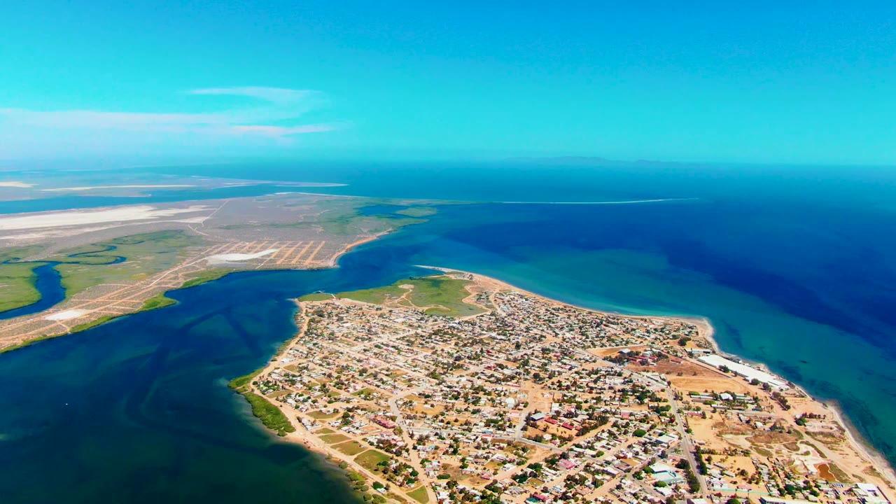Opening aerial frame over San Carlos, Baja California Sur, with the town on a sandy peninsula, blue water on both sides, distant pale flats, clear sky, and no visible people.