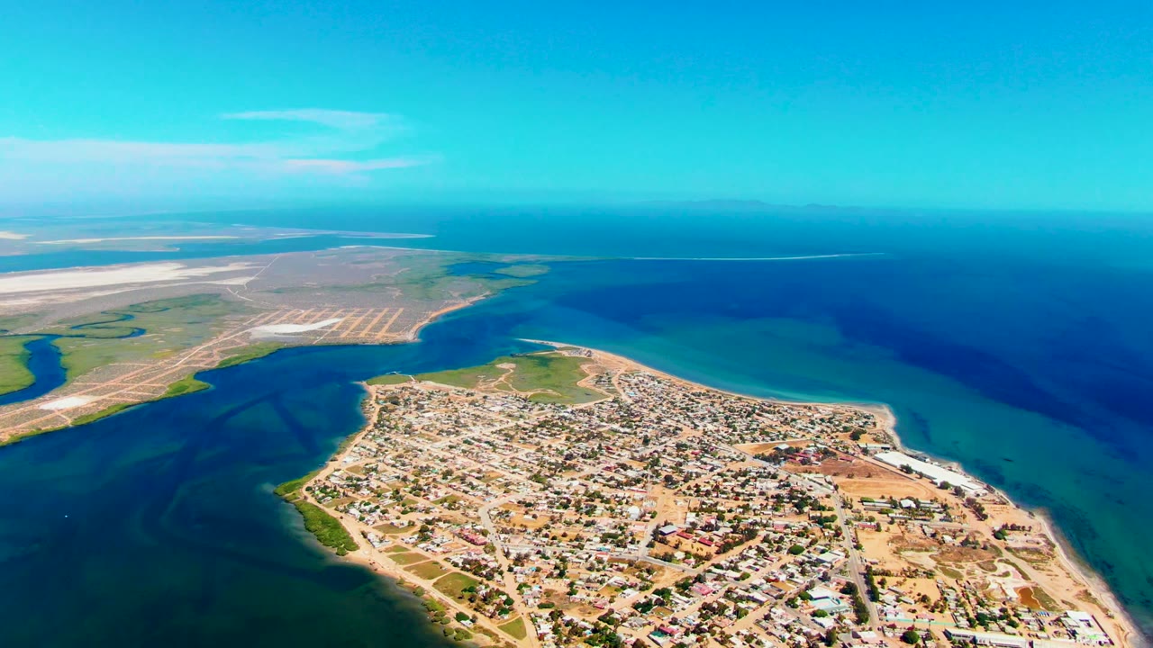 Third aerial frame over San Carlos, Baja California Sur, showing the town across the lower right, dark water at left, open blue ocean at right, pale flats in the distance, and no visible people.
