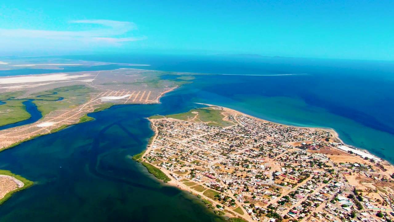 Aerial frame over San Carlos, Baja California Sur, showing a sandy coastal town beside a dark tidal inlet, turquoise shallows, deep blue ocean, pale exposed flats, and no visible people.