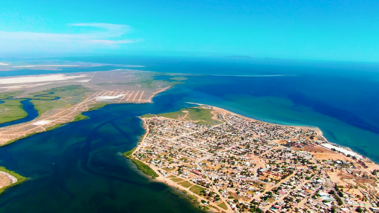Aerial view over San Carlos where the town sits along the lower right edge, the inlet darkens the foreground water, exposed flats stretch left of frame, and no visible people appear.