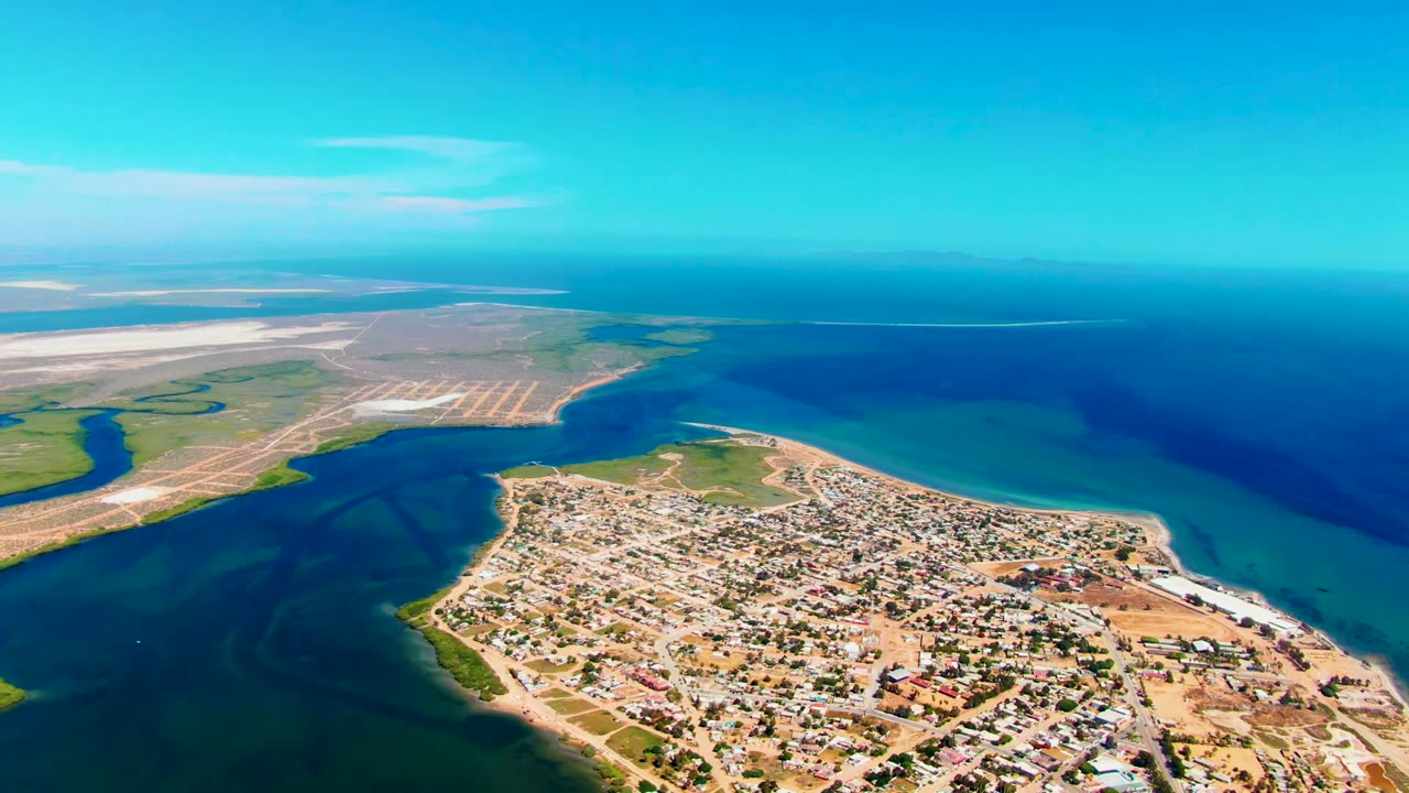 Opening aerial frame over San Carlos, Baja California Sur, showing a compact coastal town, turquoise shoreline water, darker ocean, pale flats, and no visible people.