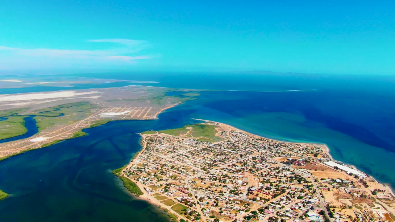 Wide aerial over town, curved shoreline, shallow water, and open ocean.