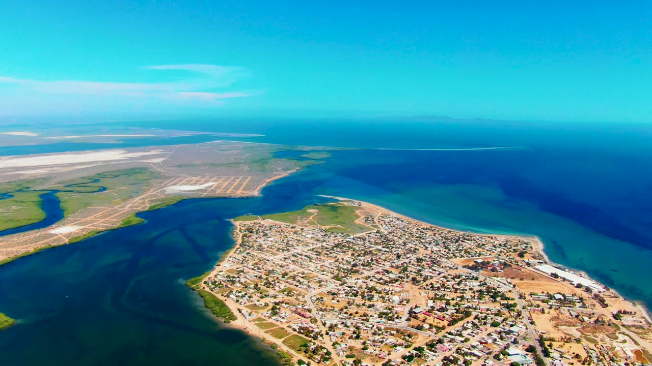 Aerial frame of San Carlos, Baja California Sur, showing dense low buildings beside blue coastal water, pale land flats in the distance, a clear sky, and no visible people.