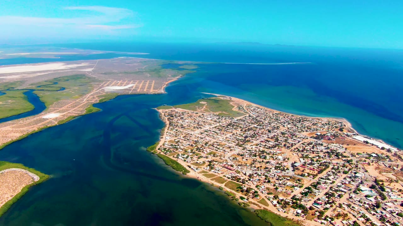 Wide aerial frame over San Carlos, Baja California Sur, with dark tidal water in front, a sandy town peninsula at center, blue ocean beyond, and no visible people.