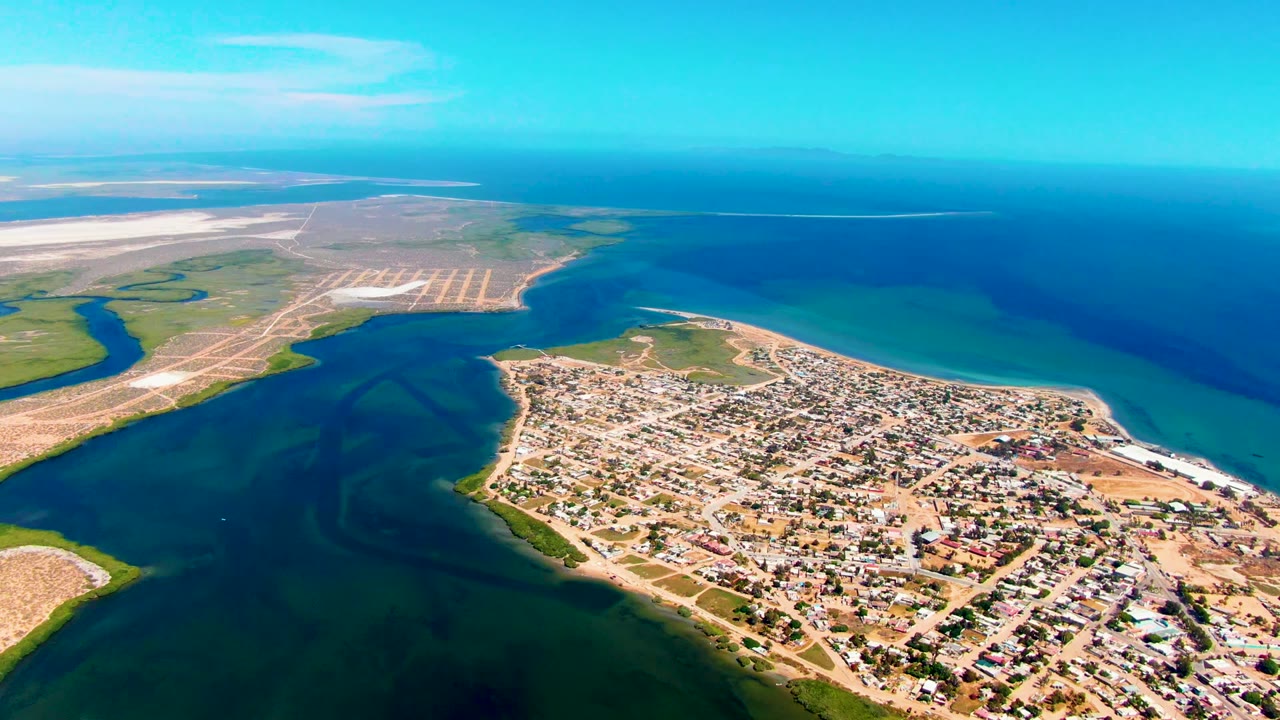 High drone frame over San Carlos, Baja California Sur, with town blocks filling the lower right, dark inlet water at left, a curved beach edge, deep blue sea at the horizon, and no visible people.