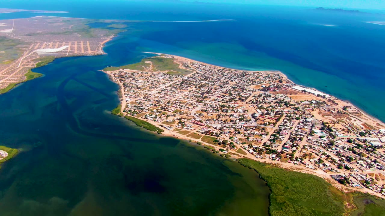 Opening aerial frame over San Carlos, Baja California Sur, showing a coastal town, inlet water in the foreground, deeper blue ocean beyond, pale patterned land at left, and no visible people.