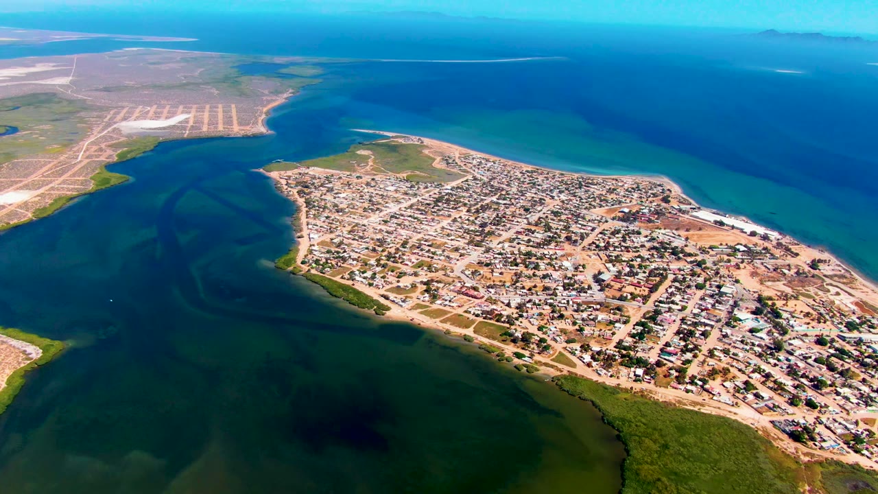 Mid-early frame over San Carlos, Baja California Sur, showing the coastal settlement across the lower right, the inlet curving through the left side, the blue ocean across the top, and no visible people.